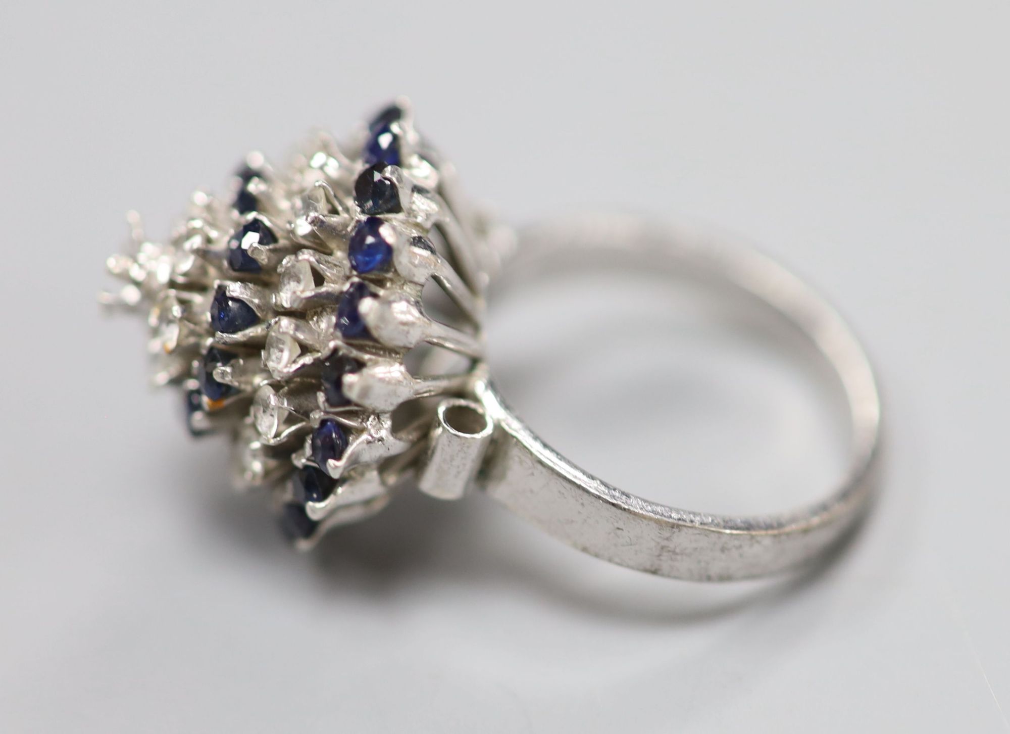 A white metal (stamped 18k) and two colour sapphire? set raised cluster ring, size K/L, gross 5.2 grams (one stone missing).